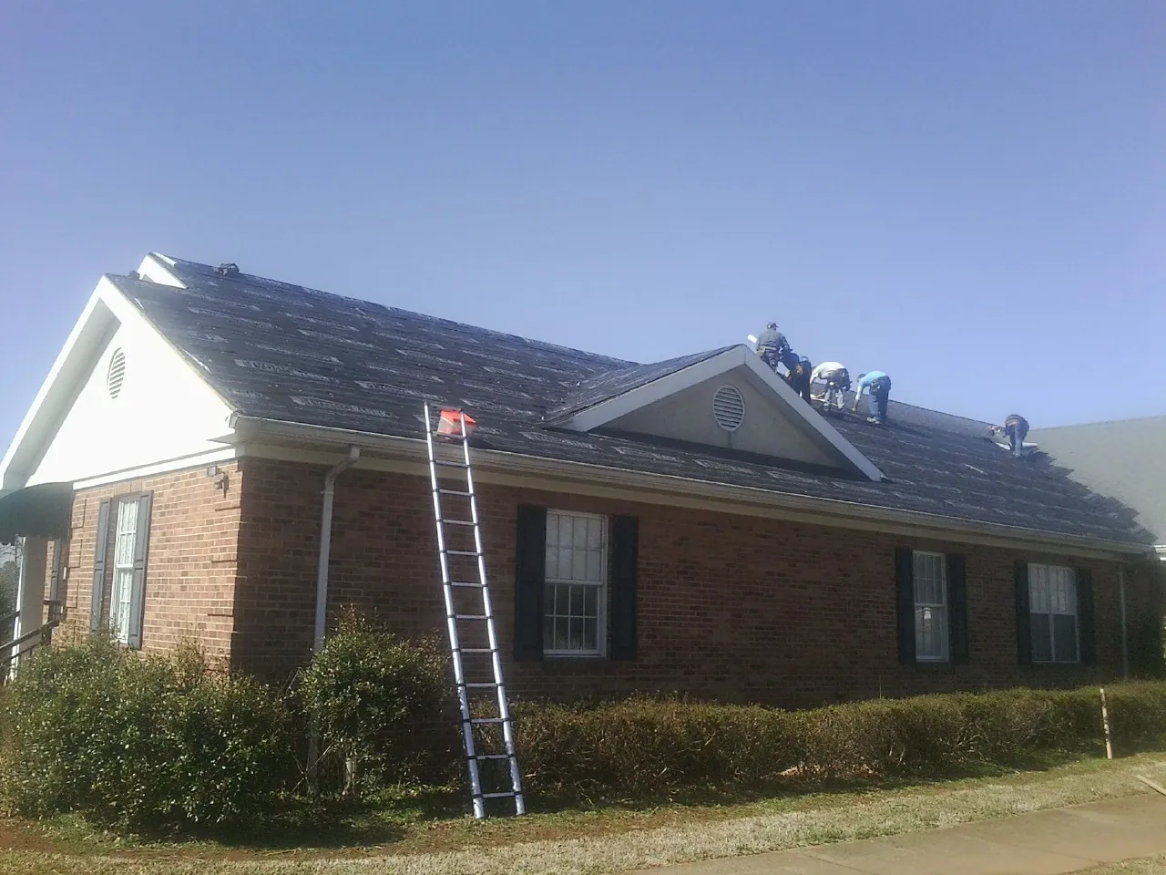 Crew tearing shingles off a brick ranch roof with a ladder set — Metal Roof Repair work by our crew in Melville